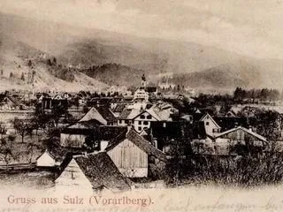 Ein altes Foto zeigt eine kleine Stadt mit traditionellen Häusern und steilen Hügeln im Hintergrund. Die Stadt liegt in Vorarlberg, Österreich.