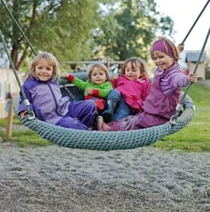 Vier junge Mädchen in Winterjacken sitzen zusammen auf einer Seilschaukel, lächeln und posieren für die Kamera. Die Schaukel steht in einem Spielplatz mit Kiesboden und grünem Rasen.