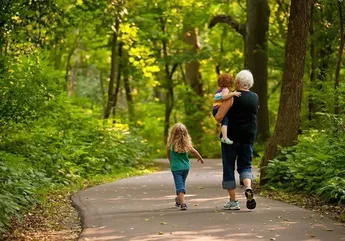 Eine ältere Frau läuft mit zwei kleinen Kindern auf einem Pfad im Wald, eines im Arm und eines neben ihr.