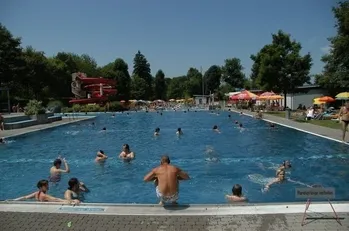 Ein großer Außenpool mit mehreren schwimmenden und sonnenbadenden Personen, umgeben von Bäumen und Sonnenschirmen. Ein Wasserrutsche ist im Hintergrund zu sehen.