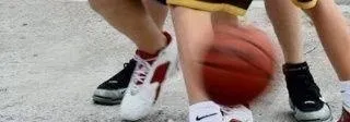 Zwei Personen spielen Basketball auf einem Platz, einer dribbelt den Ball, während der andere aufmerksam zusieht. Sie tragen Turnschuhe, ein Paar ist schwarz und weiß mit roten Akzenten, das andere komplett schwarz.