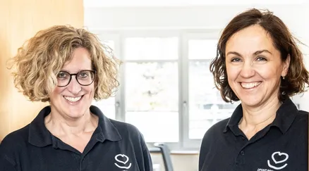 Zwei Frauen stehen nebeneinander, beide lächeln und tragen schwarze Poloshirts mit einem weißen Herzlogo auf der linken Brust. Sie stehen vor einem Fenster mit Blick auf ein Gebäude und Bäume.