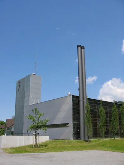 Ein modernes Gebäude mit grauer Außenseite und großen Schornsteinen oben. Das Gebäude hat viele Glasfenster und ist von grünen Bäumen umgeben.