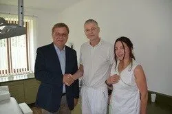 Drei Personen stehen in einem Raum. Ein Mann in Anzug und Brille schüttelt einem anderen Mann in einem weißen Hemd die Hand. Eine Frau in einem weißen Kleid steht neben ihnen. Dahinter befindet sich ein Fenster mit Jalousien und ein Waschbecken.