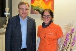 Ein Mann und eine Frau stehen nebeneinander mit einem Strauß lila Blumen davor. Sie lächeln und scheinen für ein Foto zu posieren. Hinter ihnen ist eine weiße Wand mit einem farbenfrohen Gemälde.