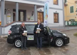 Zwei Männer stehen vor einem schwarzen Auto und halten Bücher in den Händen. Sie lächeln und scheinen für ein Foto zu posieren. Hinter ihnen steht ein Gebäude mit mehreren Fenstern und Säulen.