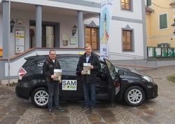 Zwei Männer stehen vor einem schwarzen Auto und halten Bücher in den Händen. Sie lächeln und scheinen für ein Foto zu posieren. Hinter ihnen steht ein Gebäude mit mehreren Fenstern und Säulen.