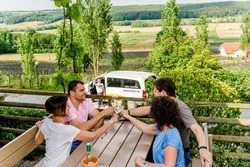 Vier Personen versammeln sich um einen Holztisch auf einem Balkon und stoßen mit Getränken an. Dahinter bietet eine ländliche Landschaft Bäume und ein entferntes Haus.