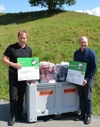 Zwei Männer stehen neben einer weißen Lagerbox voller Kleidung und halten Urkunden. Sie sind auf einer Straße mit einem grün bewachsenen Hügel im Hintergrund.