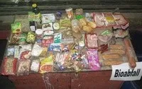 Ein Tisch ist mit verschiedenen verpackten Lebensmitteln gefüllt, darunter Würste, Käse und Brot. Es gibt ein Schild, das 'Bioabfall' besagt.