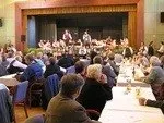 Eine Gruppe von Menschen sitzt an langen Tischen in einem Bankettsaal. Zwei Personen stehen mit Musikinstrumenten auf der Bühne. Der Saal hat Holzwände und Vorhänge.