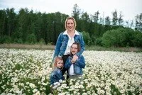 Eine Frau und zwei Kinder stehen in einem Feld weißer Blumen mit einem Wald im Hintergrund.