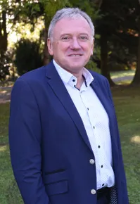 Ein Mann in einem marineblauen Blazer und einem weißen Hemd mit kleinen Punkten steht in einem Park mit grünem Rasen und Bäumen.