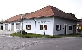 Ein weißes Gebäude mit einem roten Dach und der Aufschrift 'Bad Waltersdorf'. Es hat zwei Garagen und einen Feuerhydranten davor.