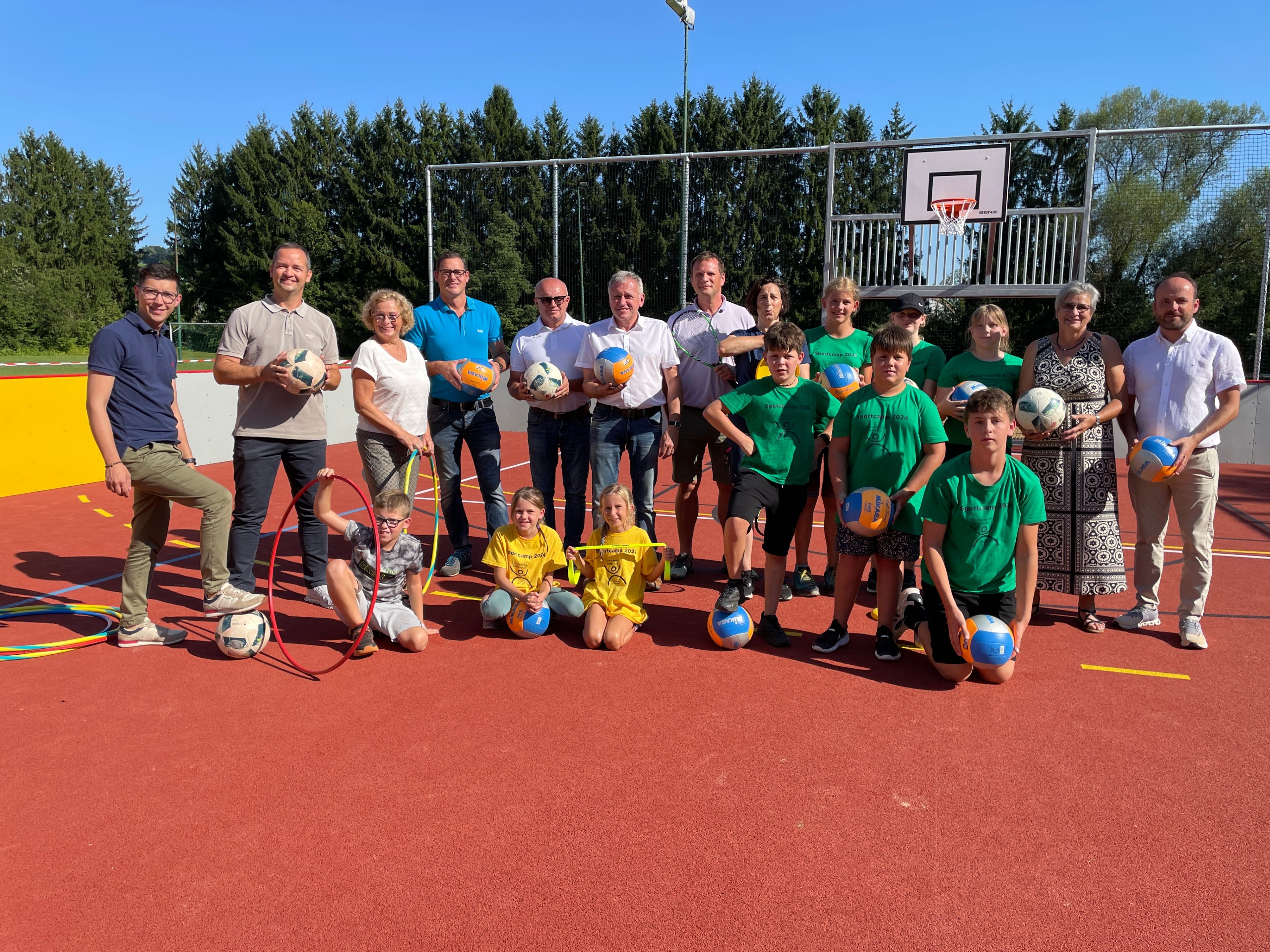 Eine Gruppe von Menschen posiert auf einem Sportplatz für ein Foto, hält verschiedene Bälle. Einige tragen grüne Hemden, andere Freizeitkleidung. Hinter ihnen ist ein Basketballkorb und Bäume zu sehen.
