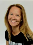 Eine Frau mit blonden Haaren lächelt und trägt ein schwarzes T-Shirt. Sie hat einen fröhlichen Gesichtsausdruck und steht vor einem schlichten Hintergrund.