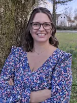 Eine Frau mit Brille und einem Blumenkleid steht lächelnd neben einem Baum mit verschränkten Armen. Hinter ihr ist ein Rasenfeld mit einem Haus.