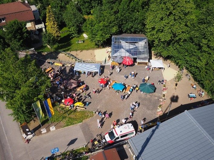  Abschnitt Bild: Eine Luftaufnahme einer Outdoor-Veranstaltung mit vielen Menschen, die sich um Tische unter bunten Sonnenschirmen versammeln, einem Gewächshaus im Hintergrund und einem roten und weißen Krankenwagen in der Nähe.