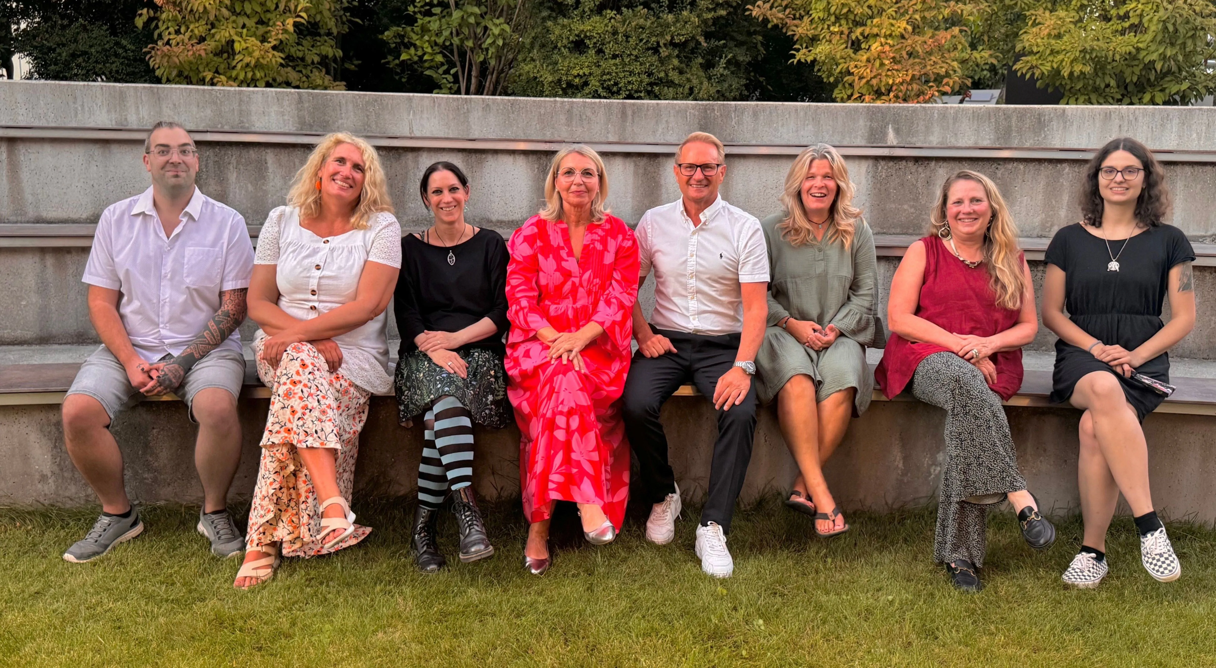 Eine Gruppe von sechs Personen, drei Frauen und drei Männer, sitzen auf einer Betonbank in einem Garten. Sie alle lächeln und scheinen für ein Foto zu posieren.