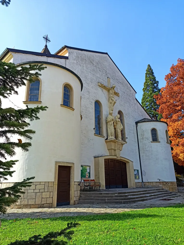 Abschnittsbild: Eine weiße Kirche mit einem Kreuz und Statuen an der Wand, zwei Rundbogenfenstern und einer Bank davor.