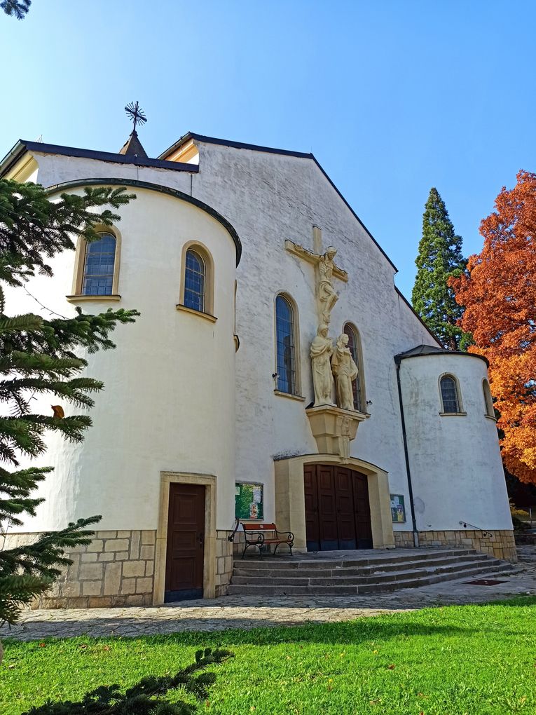Abschnittsbild: Eine weiße Kirche mit einem Kreuz und Statuen an der Wand, zwei Rundbogenfenstern und einer Bank davor.