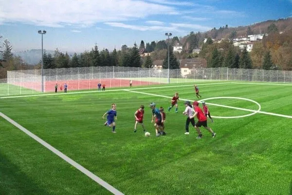 Eine Gruppe von Kindern spielt Fußball auf einem grünen Feld, umgeben von einem Zaun, mit einem Hügel und Häusern im Hintergrund.