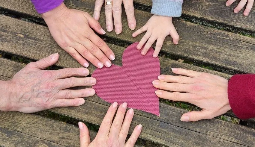 Eine Gruppe von Händen, darunter eine ältere Person, ein Kind und andere, berührt ein rosa Herz auf einer Holzbank.