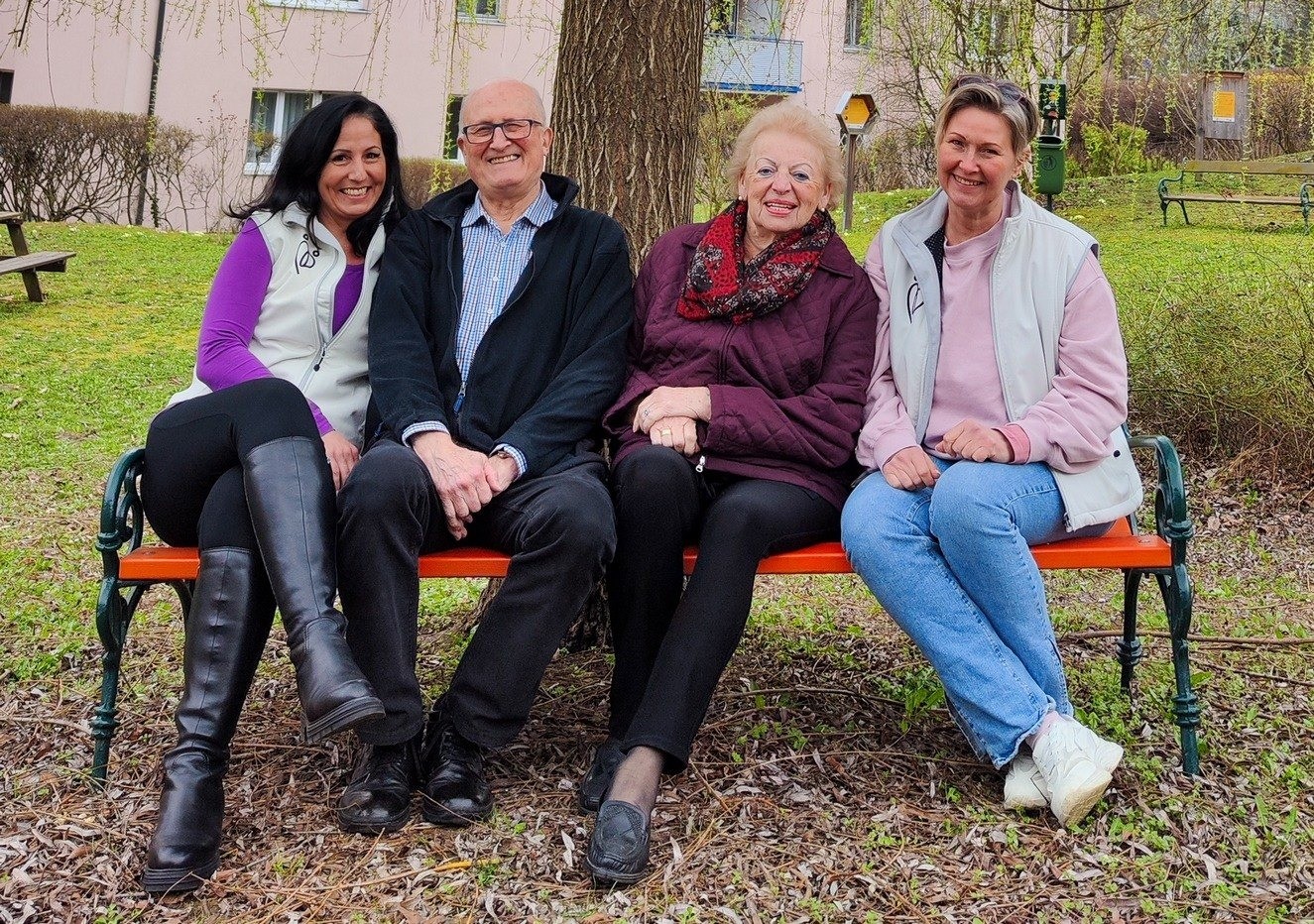 Vier Erwachsene, zwei Frauen und zwei Männer, sitzen auf einer orangefarbenen Bank in einem Park. Sie alle scheinen glücklich zu sein und lächeln für die Kamera.
