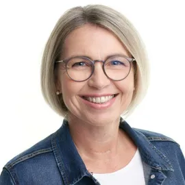 Eine Frau mit kurzen blonden Haaren, die eine Brille und eine Jeansjacke trägt, posiert lächelnd für ein Foto.