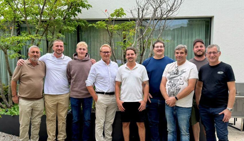 Eine Gruppe von Männern steht zusammen vor einem weißen Gebäude mit grünen Pflanzen und Bäumen. Sie lächeln und posieren für ein Foto.