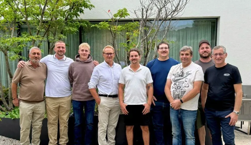 Eine Gruppe von Männern steht zusammen vor einem weißen Gebäude mit grünen Pflanzen und Bäumen. Sie lächeln und posieren für ein Foto.
