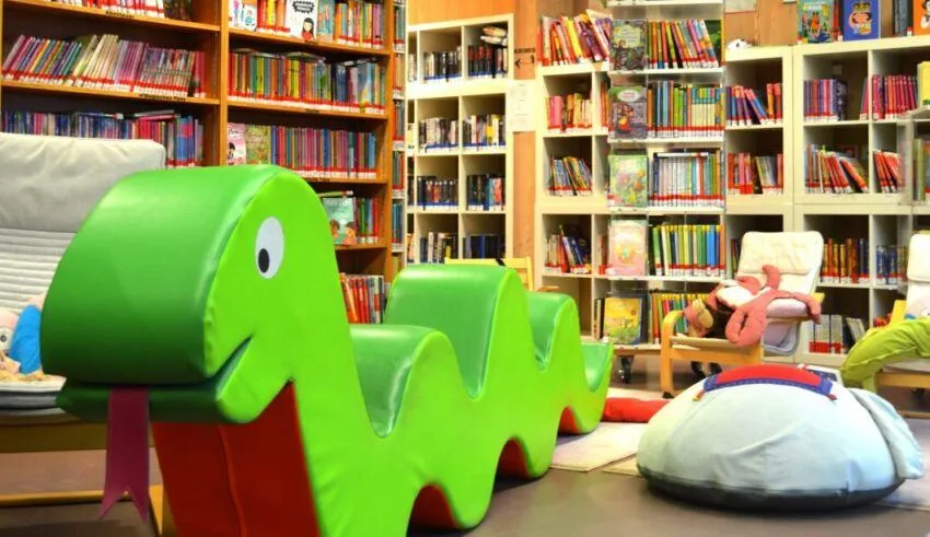Eine bunte Kinderbibliothek mit Büchern an Regalen, einem grünen Schlangenstuhl und einem Kuscheltier auf einem Stuhl.