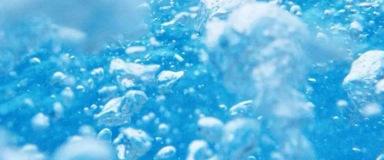 Eine Nahaufnahme von Wasser mit zahlreichen Blasen unterschiedlicher Größe, von denen einige wie Eiskristalle erscheinen, vor einem tiefblauen Hintergrund.