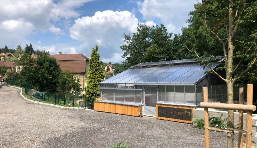 Außenansicht eines Gewächshauses mit Solarmodulen auf dem Dach. Es ist von einer Hecke und Bäumen umgeben. Ein hölzernes Schild befindet sich auf der rechten Seite.