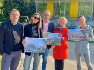 Fünf Personen stehen draußen und halten Architekturpläne in der Hand. Zwei Frauen halten große Poster, während ein Mann und zwei Frauen kleinere Pläne halten. Sie stehen vor einem gelben und weißen Gebäude mit Bäumen im Hintergrund.