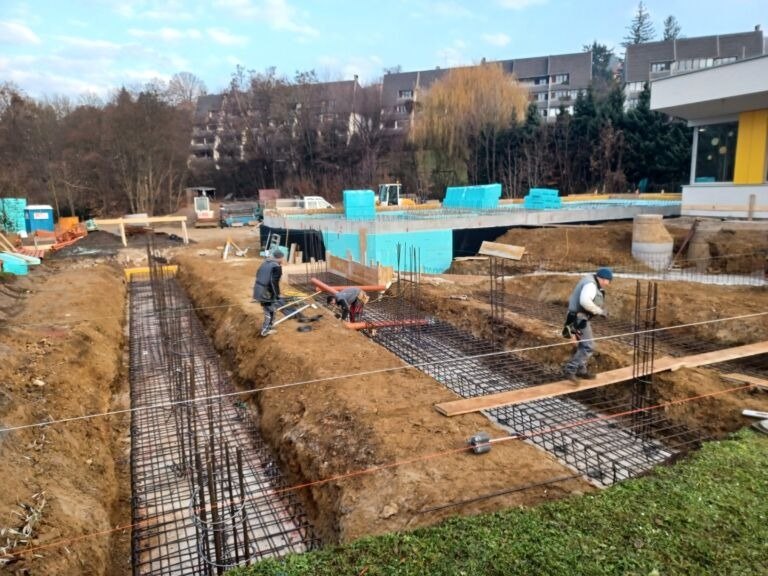 Bauarbeiter bauen eine Poolfundierung auf einer Baustelle, mit Maschinen und Gebäuden im Hintergrund.