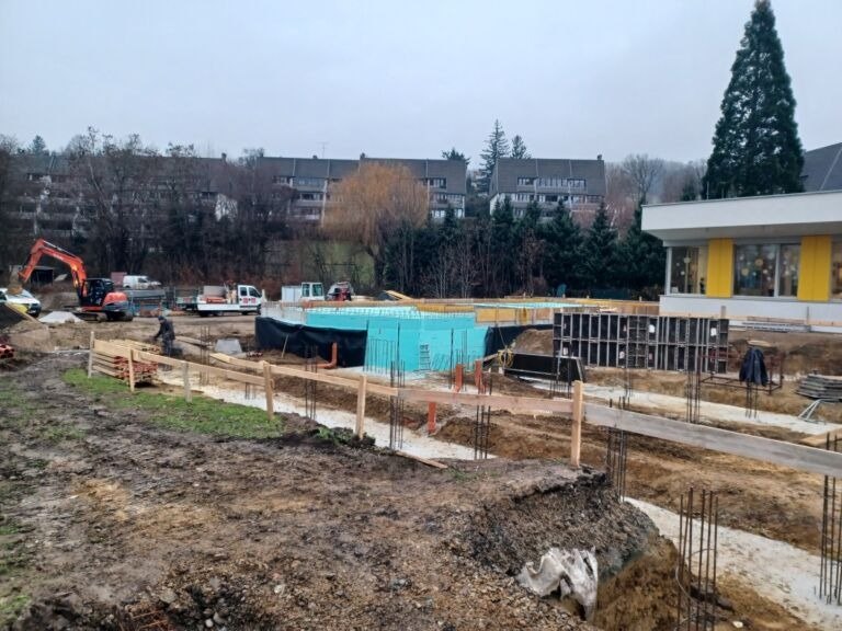 Eine Baustelle mit einem Gebäude im Bau, einer blauen Poolstruktur und verschiedenen Maschinen wie Baggern und Lastwagen. Im Hintergrund befinden sich Bäume und Wohngebäude.