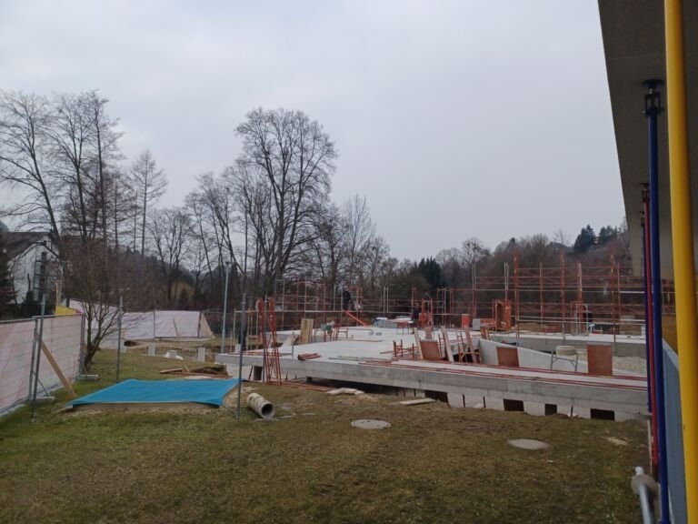 Eine Baustelle ist im Gange. Das Gelände ist von Zäunen und Gerüsten umgeben. Arbeiter sind sichtbar, und es liegen verschiedene Materialien auf dem Boden. Bäume sind im Hintergrund.
