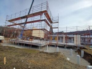 Eine Baustelle mit einem im Bau befindlichen Gebäude, das einen Stahlrahmen, Gerüst und Betonsäulen aufweist. Die Baustelle ist von Erde umgeben und hat einen klaren Himmel.