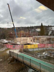 Eine Baustelle mit einem Kran, der ein Holzgebäude hebt, umgeben von Zäunen, an einem bewölkten Tag. Eine Brücke und ein Gebäude sind ebenfalls zu sehen.
