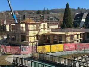 Eine Baustelle eines mehrstöckigen Gebäudes mit Gerüsten. Das Gebäude ist im Bau, einige Wände sind bereits fertig. Im Hintergrund befinden sich Bäume und Häuser.
