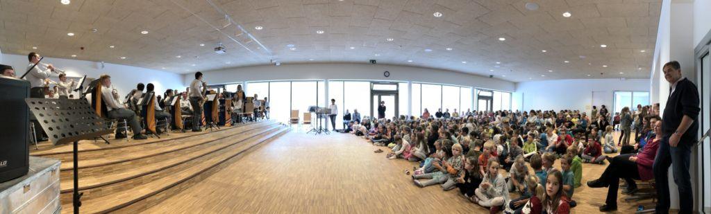 Ein großer Innenraum mit einer Band, die auf der Bühne für ein Publikum von Kindern spielt, die auf dem Boden sitzen.