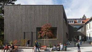 Ein modernes Gebäude mit Holzfassade steht auf einem Campus. Studenten sitzen herum, einige gehen, und ein Baum ist durch ein großes Fenster sichtbar.