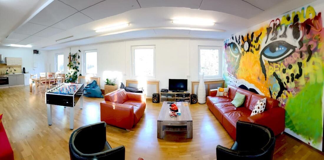 Ein Wohnzimmer mit einem großen roten Sofa, einem roten Sessel, einem Couchtisch und einem Fernseher. Es gibt auch einen Tischfußballtisch und Sitzsäcke.
