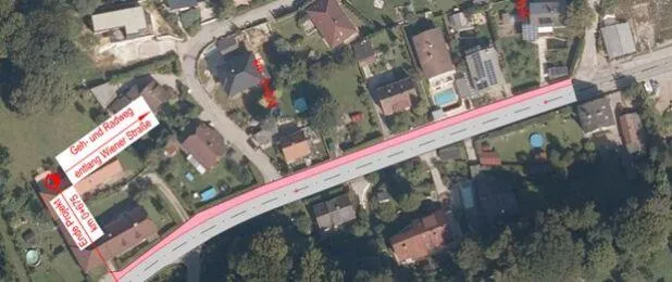 Eine Luftaufnahme eines Wohngebiets mit einer in Pink hervorgehobenen Straße, gekennzeichnet durch Bezeichnungen wie 'Gehr-und-Radweg', 'entlang Wiener Strasse' und 'Staugefahr'. Häuser, Bäume und ein Swimmingpool sind zu sehen.