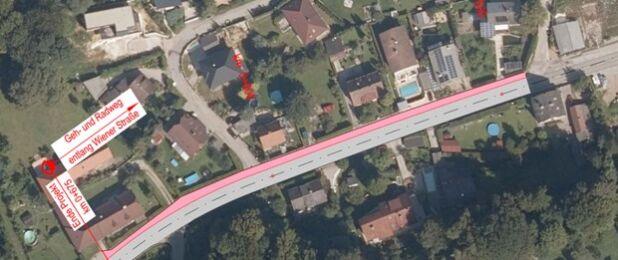 Eine Luftaufnahme eines Wohngebiets mit einer in Pink hervorgehobenen Straße, gekennzeichnet durch Bezeichnungen wie 'Gehr-und-Radweg', 'entlang Wiener Strasse' und 'Staugefahr'. Häuser, Bäume und ein Swimmingpool sind zu sehen.