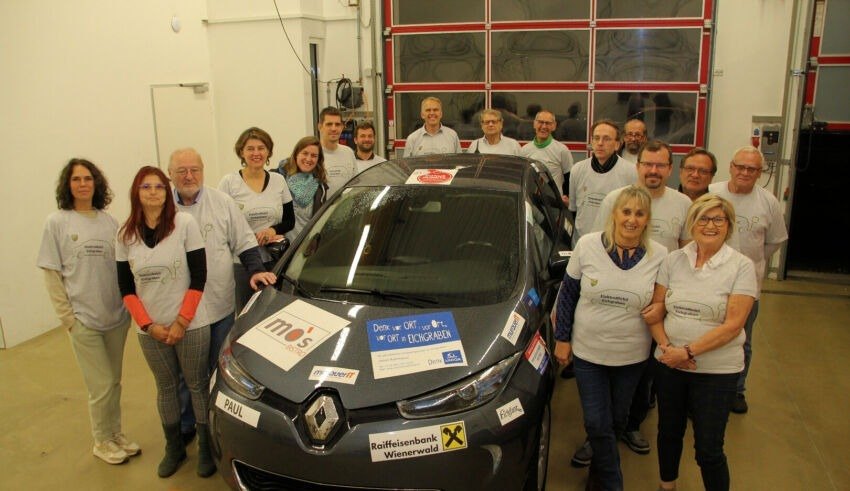 Eine Gruppe von Menschen in weißen Shirts steht lächelnd neben einem Renault Elektroauto. Einige tragen Brillen und Uhren. Auf dem Auto befinden sich Aufkleber. Hinter ihnen reflektiert eine Glastür Bilder von einem Auto und Menschen.