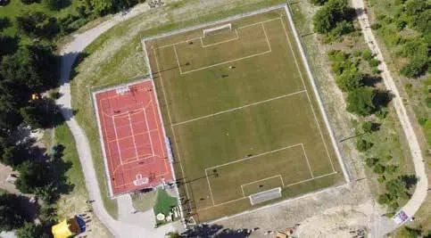 Eine Draufsicht auf ein Sportfeld mit einem roten Tennisplatz und einem grünen Fußballfeld. Das Fußballfeld hat weiße Markierungen und der Tennisplatz rote Markierungen.
