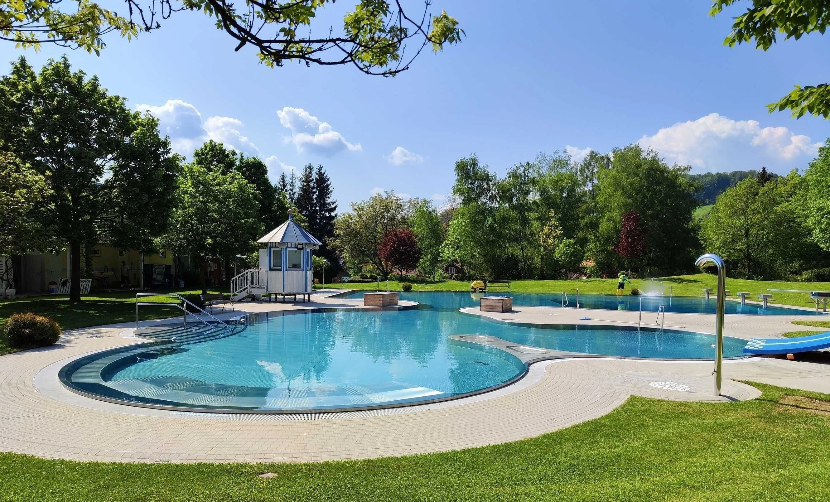 Ein großer Swimmingpool, umgeben von grünem Gras, einem kleinen Gebäude mit einer Treppe und Bäumen. Der Himmel ist blau mit einigen Wolken.