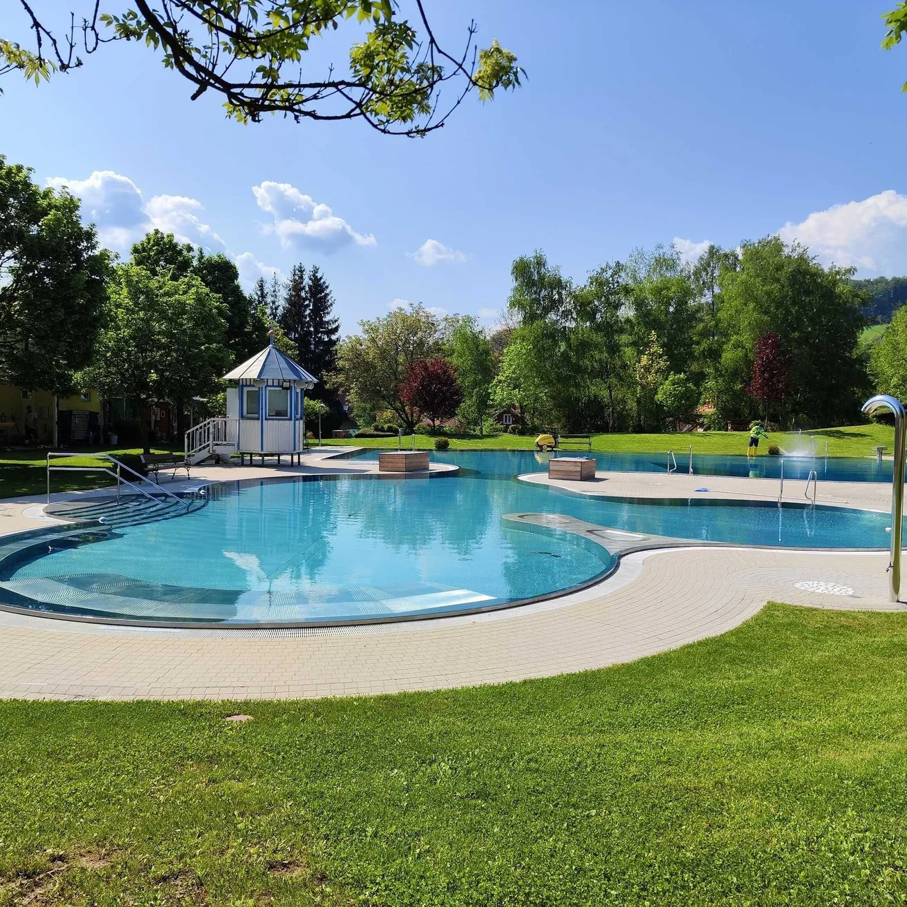 Ein großer Swimmingpool mit einem Springbrunnen befindet sich in einem Park, umgeben von grünem Gras und Bäumen. Ein kleines Pavillon steht in der Nähe, und eine Person steht neben dem Pool.